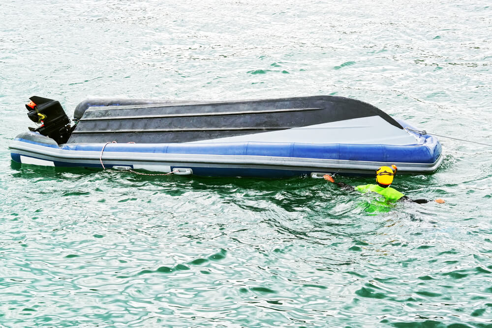 capsized inflatable boat floats on the water while a person in a bright green wetsuit and yellow helmet clings to its side